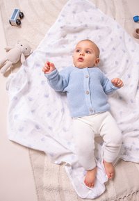 Cardigan lavorato a maglia di colore azzurro chiaro con bottoni, leggins bianchi e una coperta a fantasia. Giocattolo di peluche beige e camioncino di legno nei dintorni.