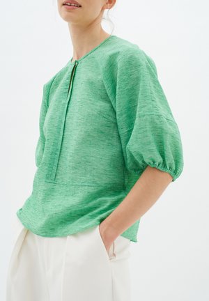 Top verde texturizzato con scollatura rotonda, chiusura frontale e maniche a sbuffo, abbinato a pantaloni color crema.