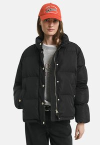 Zwarte pufferjack met een hoge kraag, grote gestikte secties, drukknoopsluitingen en een ritssluiting aan de voorkant. Gdraagt over een grijze sweatshirt en zwarte broek.