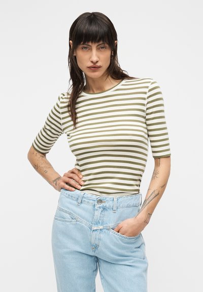 Γυναίκα με σκουρόχρωμα μαλλιά φοράει ριγέ πουκάμισο σε ελιά και άσπρο και ανοιχτόχρωμο τζιν, στέκεται με το ένα χέρι στη μέση και το άλλο στην τσέπη.