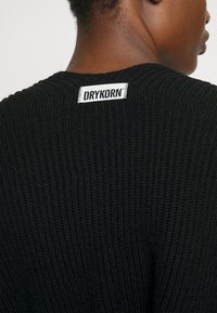 Czarny sweter z dzianiny, prezentujący strukturalny splot rib. Zawiera białą metkę z haftem "DRYKORN" umieszczoną na karku.