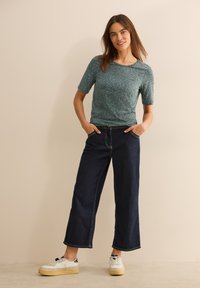 Tyrkysová tričko s krátkým rukávem a abstraktním vzorem, kombinované s tmavými volnými džínami a bílými teniskami se žlutou podrážkou, stojící proti jednolitému pozadí.