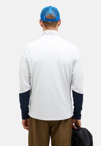 Hvid langærmet skjorte med marineblå accenter på ærmerne, med en stående krave og logo bagpå, parret med brune bukser og en sort rygsæk.