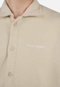 Beige button-up shirt made of textured fabric with a ribbed pattern. Features a collar and embroidered "PREDADOR" logo. Four buttons visible.