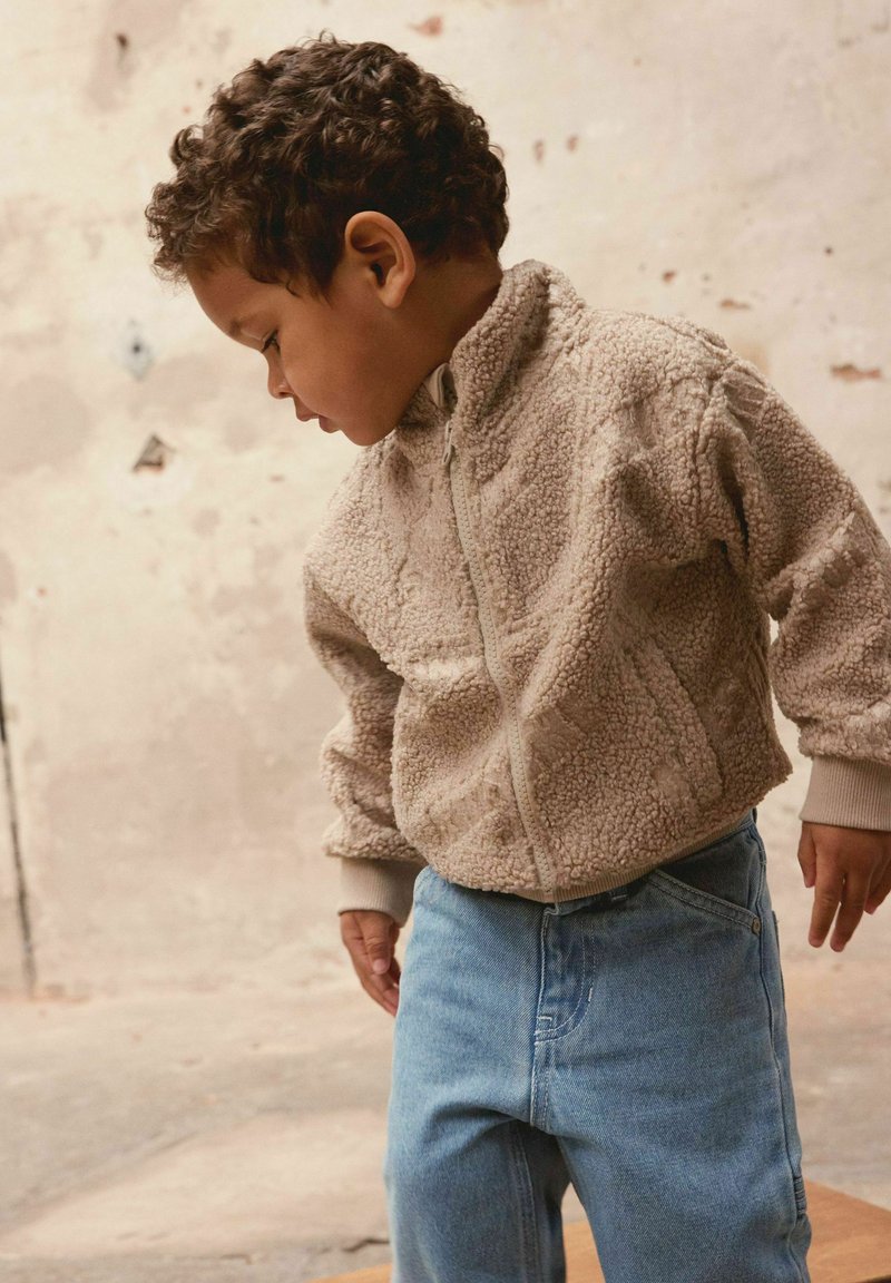 Beige fleece vest met een gestructureerd oppervlak, gecombineerd met lichtblauwe spijkerbroek. Profiel van een kind met krullend haar en handen op de heupen.