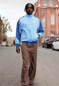 Casaco azul claro com fecho éclair e detalhes azuis, combinado com calças amplas castanhas com padrão quadriculado e sapatilhas pretas. Cenário urbano de rua.