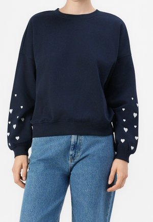 Persona que lleva una sudadera azul marino con patrones de corazones blancos en las mangas y vaqueros azul claro, de pie contra un fondo liso.