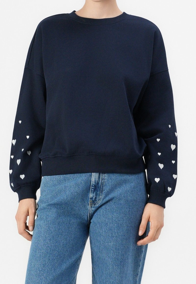 Personne portant un sweat-shirt bleu marine avec des motifs de cœurs blancs sur les manches et un jean bleu clair, debout devant un fond uni.
