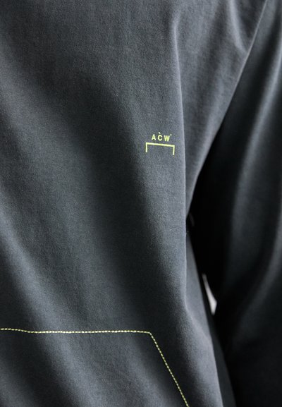 T-shirt en coton noir arborant un logo ACW en jaune vif, avec un détail de couture jaune contrastant le long de l'ourlet.