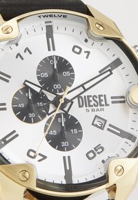 Reloj dorado con esfera blanca, subesferas negras y marcadores de hora en negro. Cuenta con una correa negra y una resistencia al agua clasificada en 5 BAR.