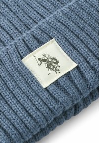 Blå, ribbestrikket stoff med en teksturert overflate, som har en hvit etikett med en svart illustrasjon av polo-spillere på hesteryggen.