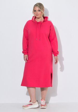 Kvinde iført en lyserød lang hættetrøjedragt med sidesplit og pink-hvide sneakers står mod en ensfarvet hvid baggrund.