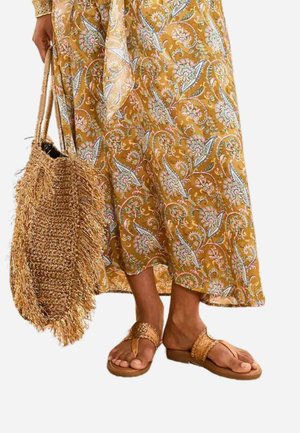 Mujer con falda floral color mostaza, sosteniendo un bolso de paja tejido y llevando sandalias entrecruzadas a juego en tonos naturales.