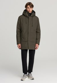 Parka verde oliva con cappuccio, coulisse e zip frontale. Indossato con pantaloni neri e sneakers grigie. Tessuto liscio, lunghezza al ginocchio.