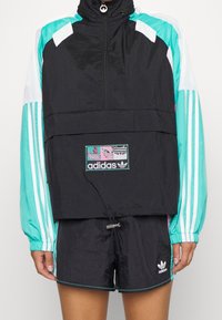 Černá a modrozelená větrovka s polovičním zipem, elastickými manžetami a logem. Doplněno černými šortkami se švem po stranách.