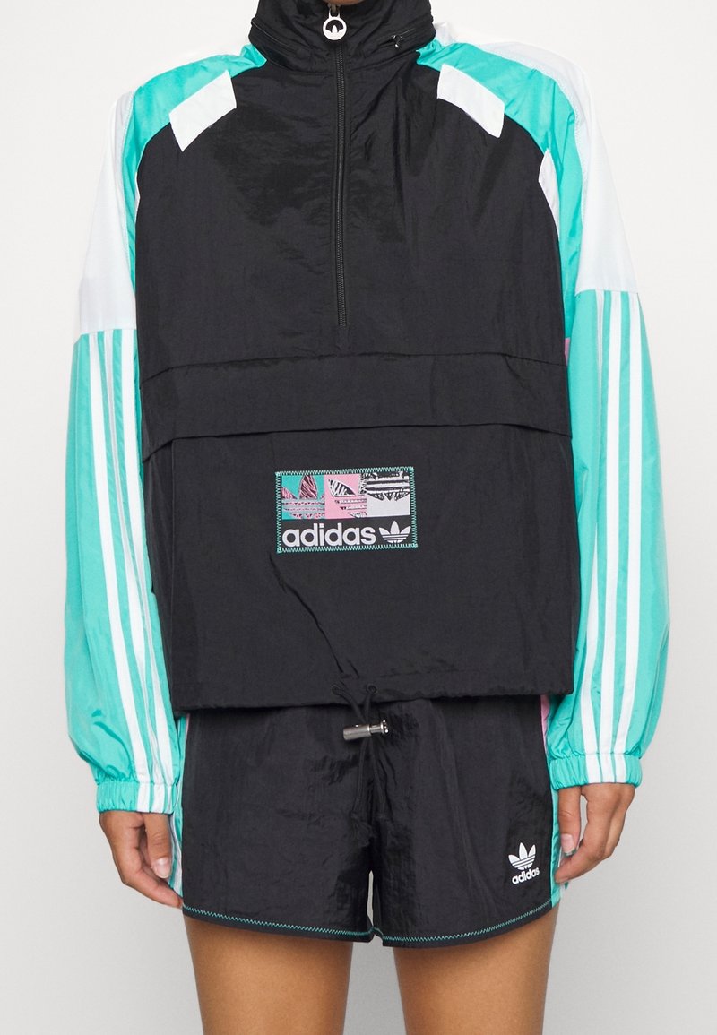Černá a modrozelená větrovka s polovičním zipem, elastickými manžetami a logem. Doplněno černými šortkami se švem po stranách.