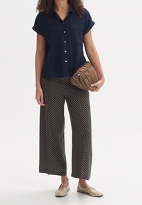 Kobieta w granatowej koszuli z krótkim rękawem na guziki, wzorzystych szerokich spodniach, beżowych butach, trzymająca brązową pikowaną torebkę kopertówkę.