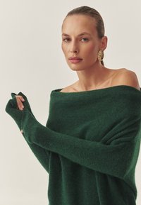 Roheline kudumiga sviiter avatud õlavarre disainiga, pikad varrukad, tekstuuriga kangas ja sõrmeaugud, mida täiustavad suured kuldsed kõrvarõngad.