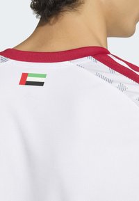 Maillot de sport blanc avec un col rouge, orné d'un petit écusson représentant un drapeau aux couleurs rouge, vert, blanc et noir au dos.