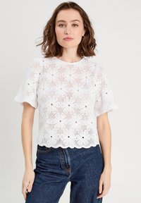 Weißes Spitzentop mit Blumenmuster, kurzen Ärmeln und gewelltem Saum, kombiniert mit dunklen Jeans. Leichtes Material, texturiertes Design.