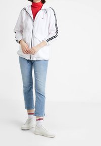 Biała lekka kurtka z czarnymi pasekami po bokach, zapinana na zamek, z kapturem. Czerwony golf, jasnoniebieskie dżinsy oraz białe trampki z teksturowanymi akcentami.