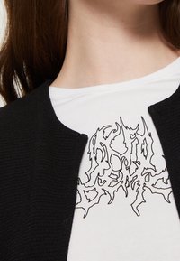 Czarny kardigan o fakturze, noszony na białej koszuli z nadrukiem w kolorze czarnym, przedstawiającym skomplikowane, postrzępione wzory linii.