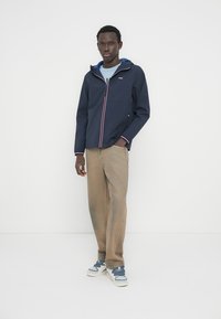 Μπλε ναυτικό τζάκετ με φερμουάρ, με αντιθετικές κόκκινες και λευκές ρίγες, ελαφρύ ύφασμα, κουκούλα προσαρτημένη και δύο πλαϊνές τσέπες, σε συνδυασμό με μπεζ παντελόνι και αθλητικά παπούτσια.