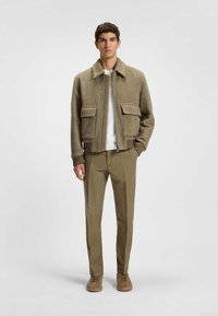 Veste en cuir brun texturé avec deux poches avant, chemise blanche en dessous, pantalon kaki et chaussures brunes. La veste a un col et des poignets côtelés.