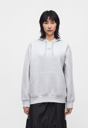 Junge Frau mit schwarzen Haaren, die einen hellgrauen Adidas-Hoodie und einen schwarzen Rock trägt, steht vor einem schlichten weißen Hintergrund.
