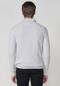 Grijze gebreide trui met een kraag en geribbelde mouwen en onderkant, gecombineerd met zwarte broek, gezien van achteren.