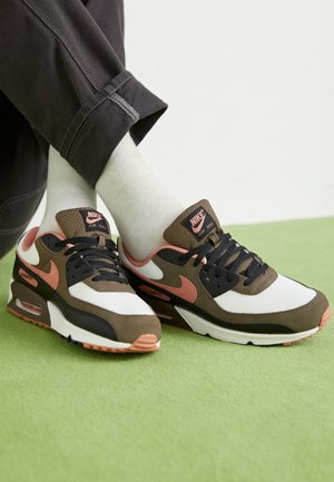 Henkilö, joka istuu ristissä jaloin vihreällä pinnalla, valkoiset sukat jalassa ja jalassa ruskeat, mustat, valkoiset ja persikanväriset Nike Air Max -lenkkarit.