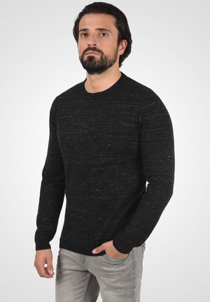 Zwart textuurtrui met lange mouwen en een ronde hals, gedragen over grijze jeans. Subtiele lichte vlekken voegen visuele interesse toe.