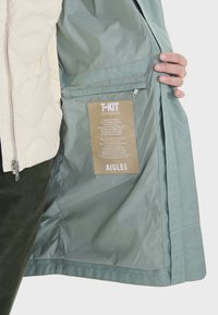 Veste parka avec une coque extérieure crème à texture matelassée, une doublure intérieure lisse vert clair et une étiquette indiquant les instructions d'entretien.