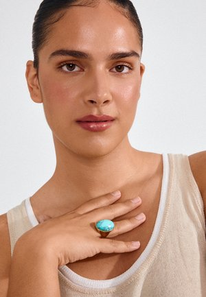 Mujer con piel suave y cabello recogido lleva una camiseta sin mangas beige y un gran anillo de oro con una piedra turquesa en su dedo cerca de la clavícula.