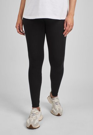 Személy fekete leggingset, bézs részletekkel ellátott fehér sportcipőt és fehér felsőt visel, egy egyszerű világos háttér előtt áll.