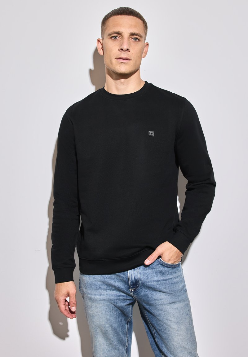 Schwarzer Sweatshirt mit Rundhalsausschnitt, langen Ärmeln und texturiertem Stoff; mit einem kleinen Logo auf der Brust. Kombiniert mit hellblauen Jeans.