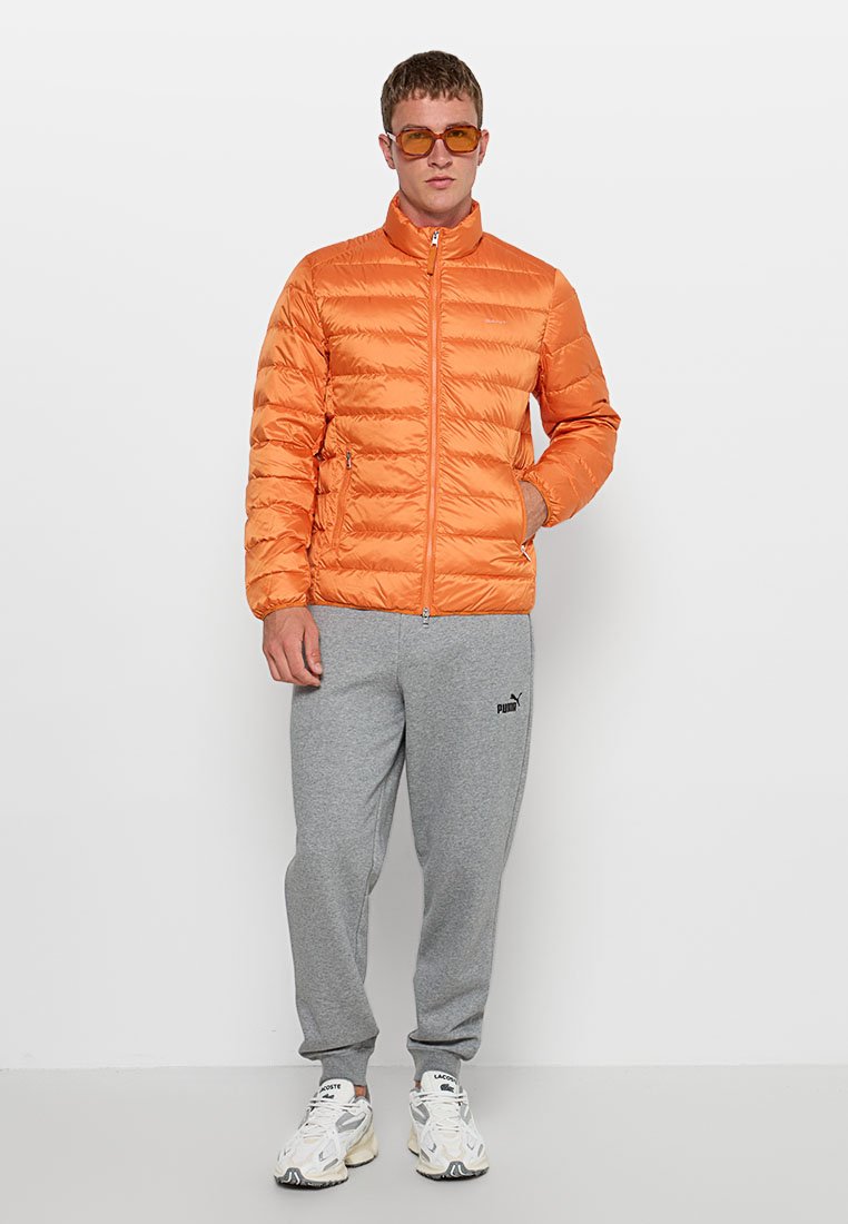 Orangene Puffjacke mit hohem Kragen und gestepptem Design, kombiniert mit grauen Jogginghosen. Bemerkenswerte Details sind die Reißverschlusstaschen.