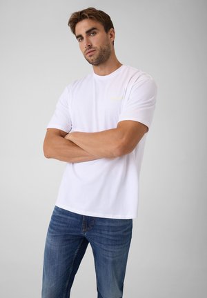 Homme aux cheveux courts et bruns, portant un t-shirt blanc et un jean bleu, debout les bras croisés devant un fond gris uni.