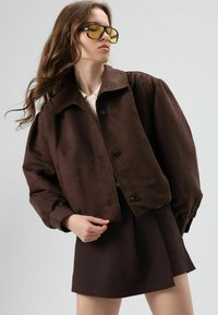 Bruine cropped jas met een kraag, knoopsluiting en pofmouwen. Draagt over een donkere rok, gecombineerd met geelgetinte zonnebril.