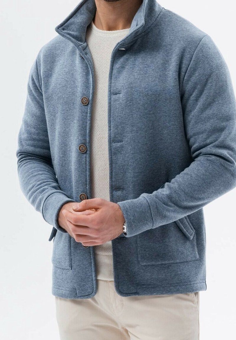 Homme portant une veste polaire bleue boutonnée sur une chemise blanche texturée avec un pantalon beige, les mains jointes devant.