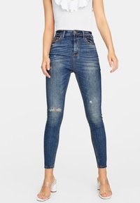 Höga, blå denimjeans med slim fit, med slitna detaljer vid knäna och kontrasterande sömmar. Matchas med genomskinliga klacksandaler.