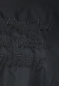 Un tissu noir présente un texte en relief dans une police dentelée indiquant : « JE TE HANTE DANS TON SOMMEIL ET LE FUTUR. » La texture semble lisse.