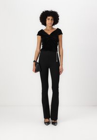 Top de terciopelo negro con escote retorcido, combinado con pantalones de tiro alto de campana negros y tacones de punta negra. Pequeño bolso negro con cadena.