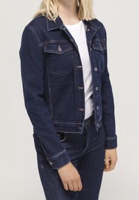 Mörkblå denimjacka med knäppning fram, två bröstfickor och klassisk krage. Har kontrasterande orange sömmar och metallknappar.