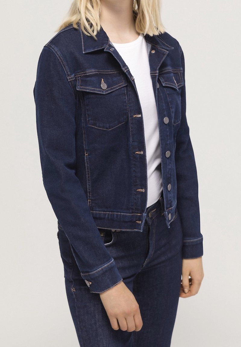 Mörkblå denimjacka med knäppning fram, två bröstfickor och klassisk krage. Har kontrasterande orange sömmar och metallknappar.