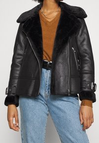 Femme portant une veste en cuir avec col en shearling noir, un pull V en laine marron, un jeans bleu clair et un collier en chaînes argentées.