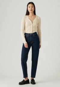 Geribbelde crèmekleurige cardigan met V-hals en knopen, gecombineerd met donkerblauwe hoge taille jeans en zwarte instappers. Glad materiaal, aansluitend ontwerp.
