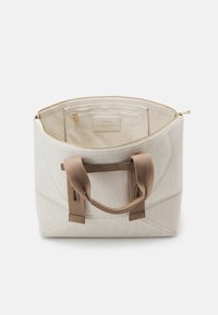 Beige stoffen draagtas met een gestructureerd ontwerp, voorzien van een ritssluiting, twee bruine canvas handvatten en een binnenzak met een label.