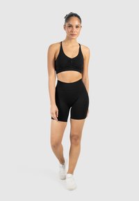 Een jonge vrouw staat zelfverzekerd in zwarte sportkleding, met een aansluitend crop-top en shorts, en witte sneakers op een neutrale achtergrond.