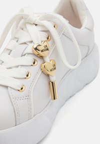 Fehér sneaker sima bőr felsőrésszel, aranyszínű, szív alakú fűzőlyukakkal, fehér fűzőkkel és texturált gumitalppal. Kiemelkedő "LOVE MOSCHINO" részlet.